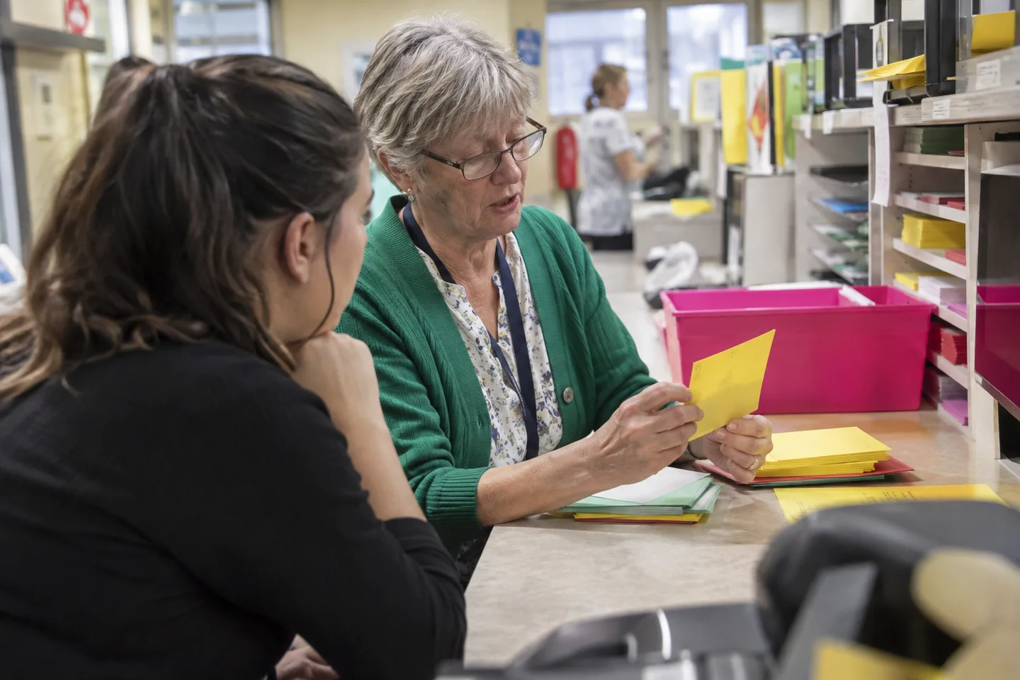 An experienced coordinator explaining a request card to a colleague at a service counter